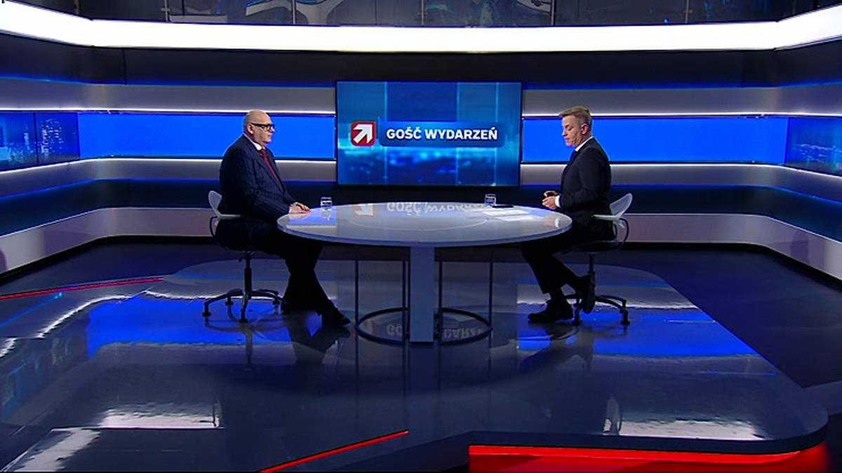 Dw&oacute;ch mężczyzn w garniturach siedzi przy okrągłym stole w studiu telewizyjnym.