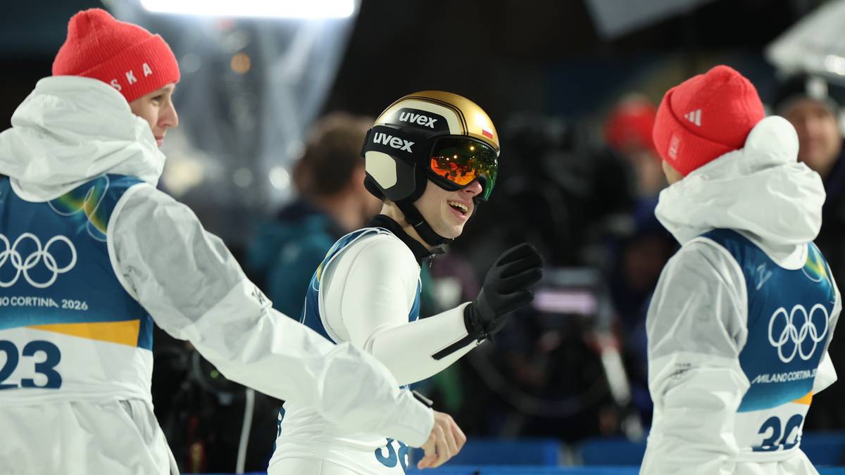 Trzech zawodników w strojach narciarskich, dwóch w białych kombinezonach i czerwonych czapkach, jeden w białym stroju i złoto-czarnym kasku, uśmiecha się i rozmawia.