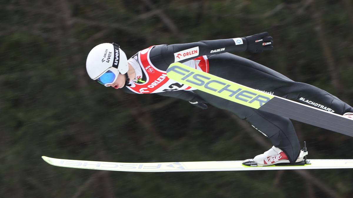 Skoczek narciarski w czarnym kombinezonie i białym kasku podczas lotu na nartach.