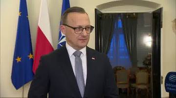 Mężczyzna w garniturze i okularach na tle flag Polski i Unii Europejskiej.