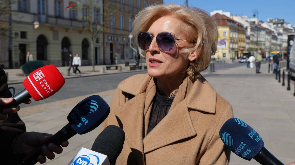 Kobieta w okularach przeciwsłonecznych i płaszczu przemawia do mikrofon&oacute;w kilku redakcji.