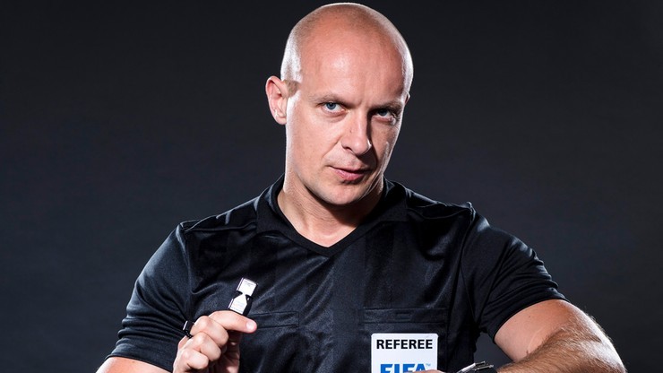 Szymon Marciniak, sędzia piłkarski w czarnej koszulce z napisem "REFEREE" i logo FIFA, trzymający gwizdek.