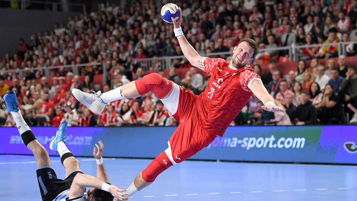 Piłkarz ręczny w czerwonym stroju wykonuje rzut do bramki, drugi zawodnik leży na parkiecie.