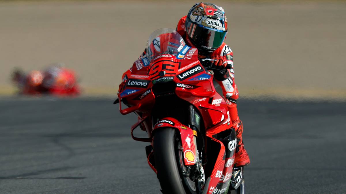 Kierowca motocyklowy w czerwonym kombinezonie i kasku jedzie na motocyklu po torze wyścigowym. W tle widać innego motocyklistę.