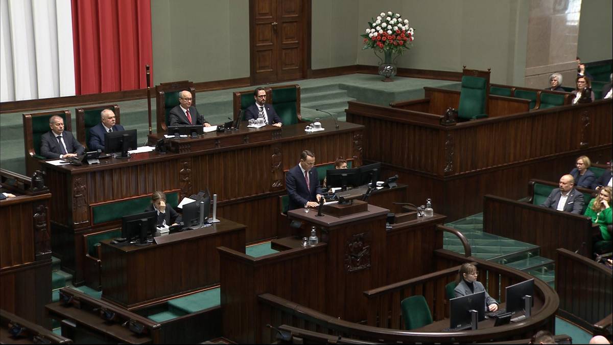 Wnętrze sali sejmowej z drewnianymi ławami i mównicą, na której stoi osoba przemawiająca.