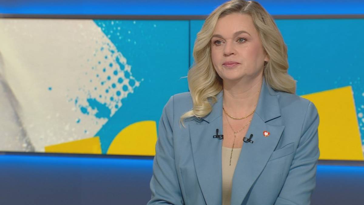 Barbara Nowacka w jasnoniebieskiej marynarce siedzi w studiu telewizyjnym z kolorowym tłem.