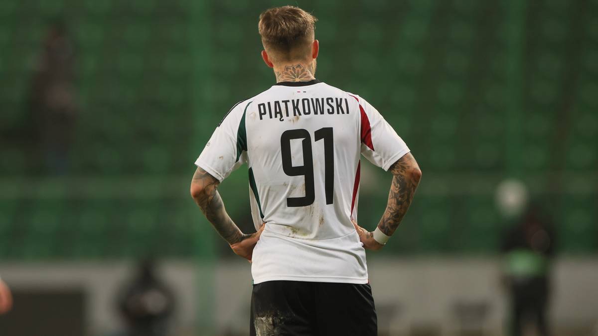 Piłkarz w białej koszulce z numerem 91 i nazwiskiem Piątkowski na plecach stoi na boisku piłkarskim.