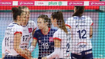 Cztery siatkarki w białych strojach sportowych stoją blisko siebie, otoczone siatką, z widocznymi numerami na koszulkach.