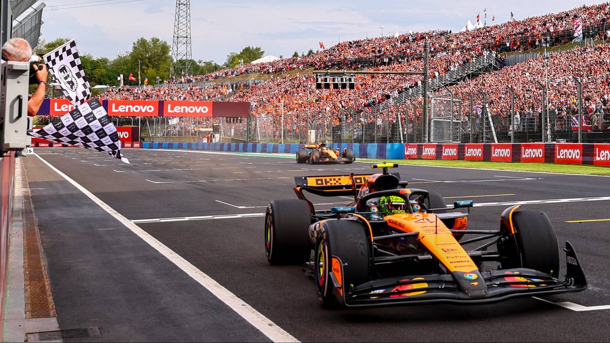 Samochód Formuły 1 zespołu McLaren z numerem 4 wyjeżdża na metę, za nim drugi samochód McLarena. Na trybunach zasiadają liczni kibice, a flaga w szachownicę jest machana przez obsługę toru.