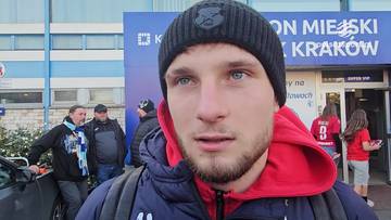 Kacper Duda, piłkarz, w czapce zimowej i bluzie z kapturem, na tle stadionu miejskiego w Krakowie.
