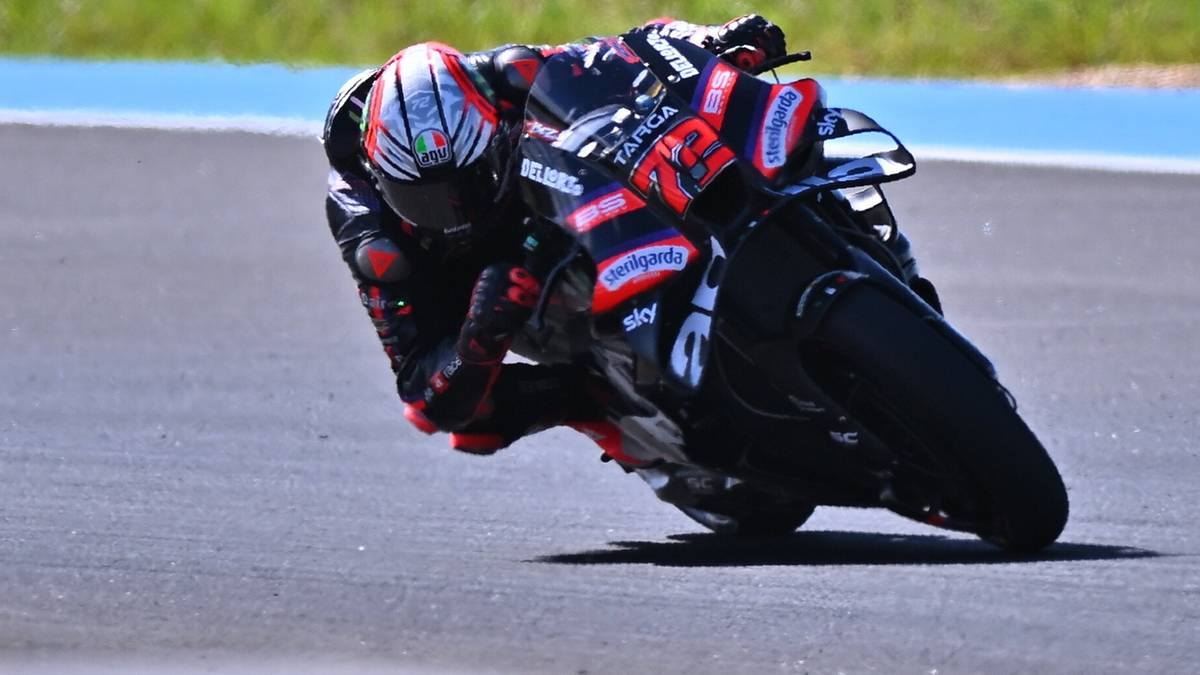 Motocyklista w czerwono-czarnym kombinezonie z białym kaskiem pochyla się mocno w zakręcie na torze wyścigowym.