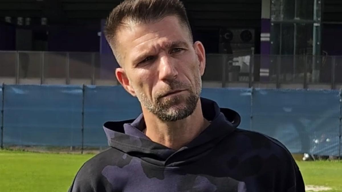 Mężczyzna z kr&oacute;tką, ciemną brodą i włosami, ubrany w bluzę z kapturem z wzorem kamuflażu.