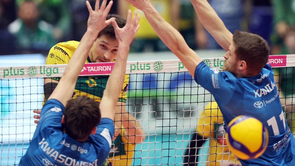 Dw&oacute;ch siatkarzy w niebieskich strojach pr&oacute;buje zablokować atak przeciwnika przy siatce podczas meczu siatk&oacute;wki.