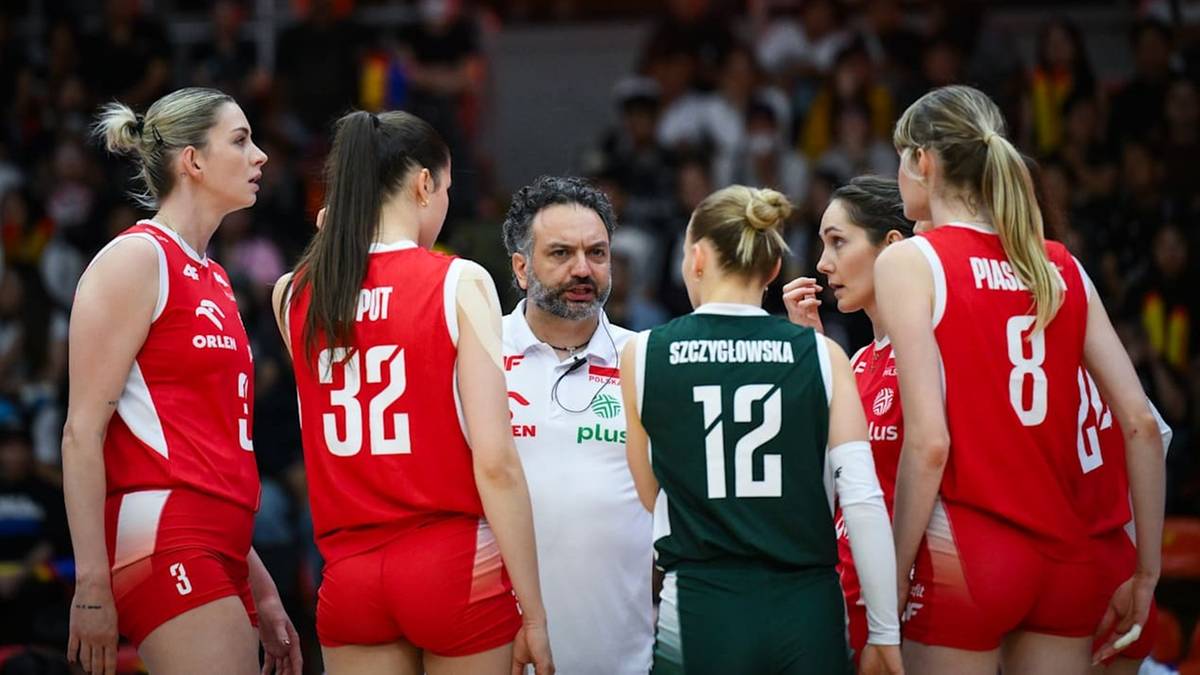 Siatkarki w czerwonych strojach otaczają trenera, słuchając go podczas przerwy w meczu.