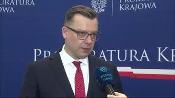 Mężczyzna w czarnym garniturze i czerwonym krawacie, udzielający wypowiedzi przed tablicą z napisem "Prokuratura Krajowa". Trzyma mikrofon z logo Polsat News.