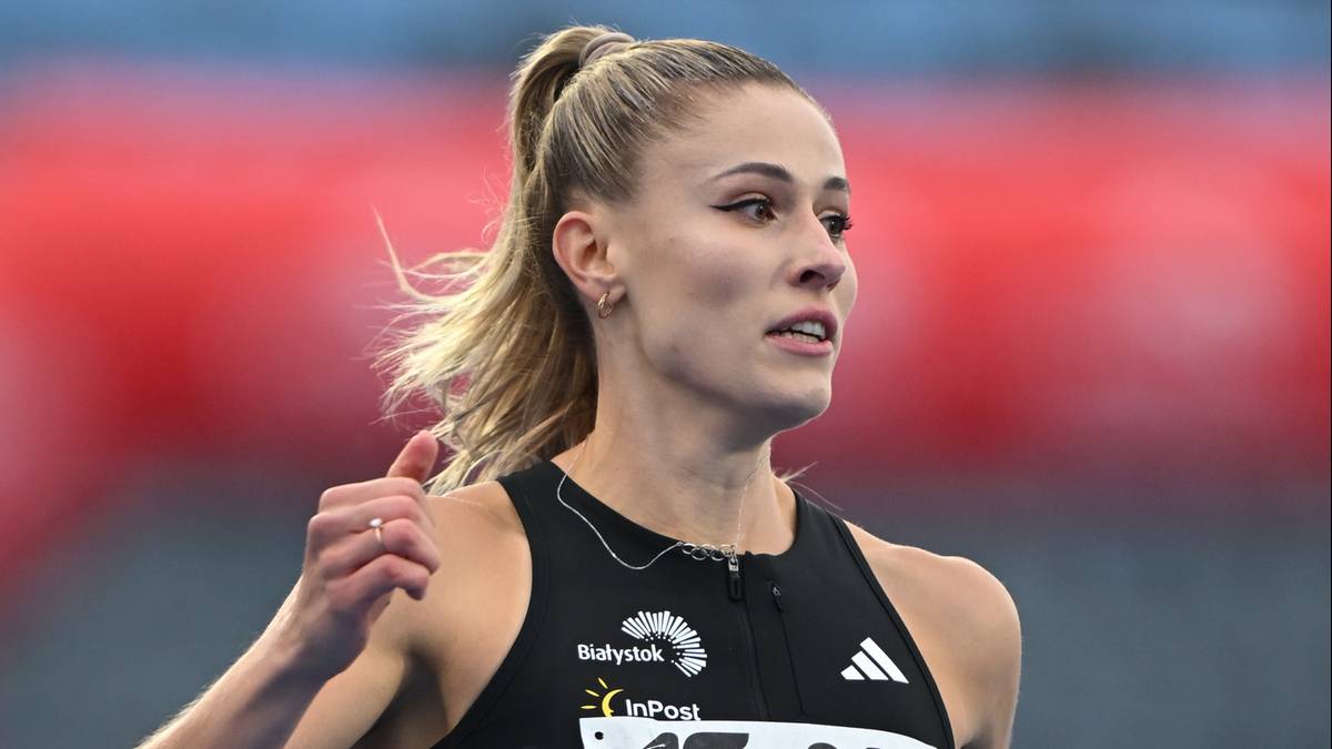 Zawodniczka lekkoatletyczna w czarnym stroju sportowym z widocznym logo sponsor&oacute;w na klatce piersiowej, z uniesionym kciukiem prawej dłoni, w tle rozmyty czerwony obiekt.