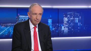 Starszy mężczyzna w garniturze i czerwonym krawacie, siedzący przed ekranem z nocnym widokiem miasta.