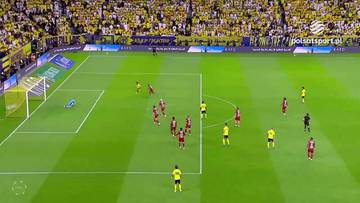 Boisko piłkarskie widziane z góry, z zawodnikami w żółtych i czerwonych strojach w trakcie meczu. Widoczne są bramki, trybuny z kibicami w tle oraz fragment murawy.