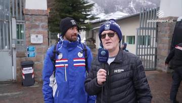 Dw&oacute;ch mężczyzn stoi przed budynkiem. Jeden z nich, w niebieskiej kurtce z białymi i czerwonymi pasami oraz czapce z pomponem, trzyma w ręku mikrofon. Drugi, w czarnej kurtce i niebieskiej czapce, patrzy w stronę obiektywu.