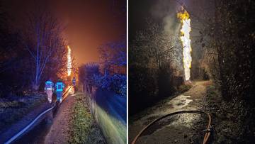 Wysoki słup ognia płonący nocą, z widocznymi sylwetkami strażaków i wężem strażackim w pobliżu.