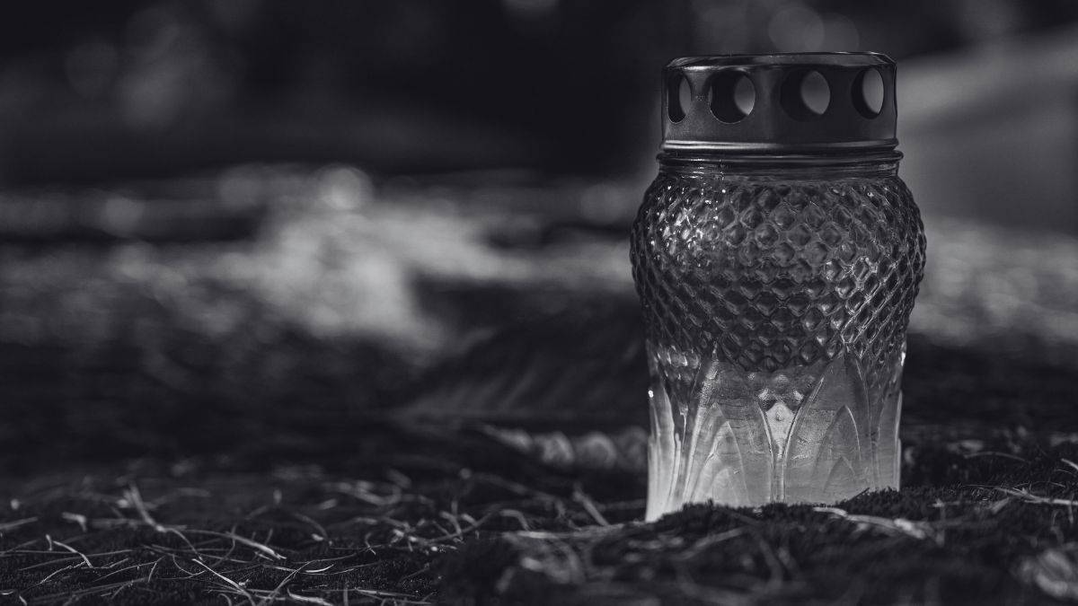 Szklany znicz z ozdobnym wzorem, stojący na ziemi pokrytej igliwiem.