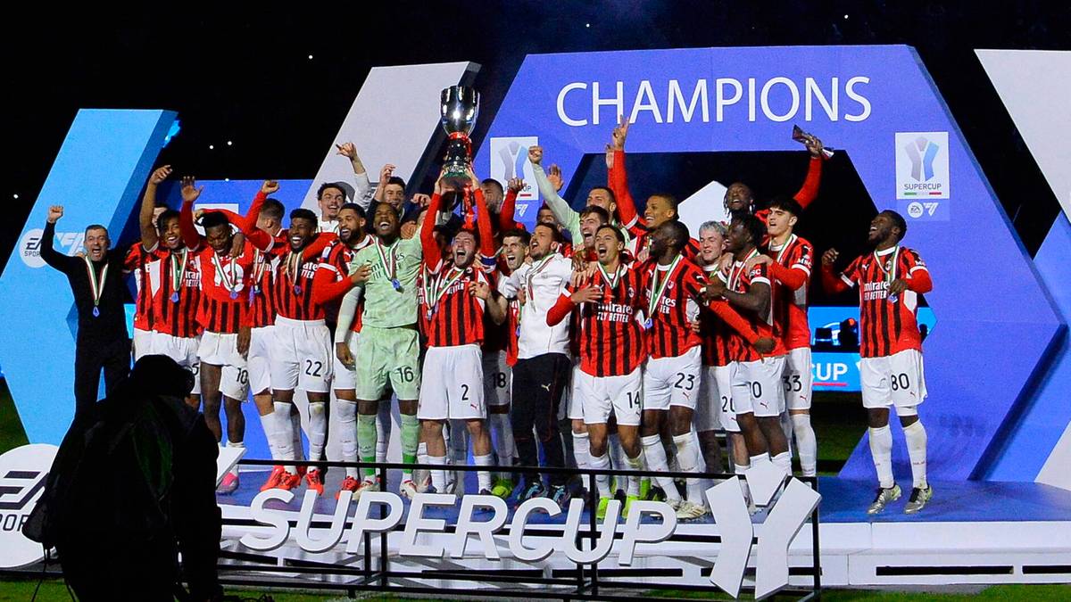 Drużyna piłkarska w czerwono-białych strojach świętuje zwycięstwo z pucharem w rękach na tle niebieskiego napisu "CHAMPIONS" i "SUPER".