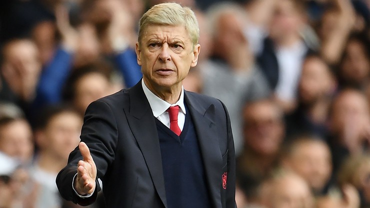 Arsene Wenger, trener piłkarski, w garniturze i krawacie, gestykuluje prawą ręką, w tle trybuny stadionu.