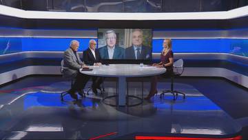 Trzy osoby siedzą przy okrągłym stole w studiu telewizyjnym, naprzeciwko nich na dużym ekranie wyświetlany jest obraz dw&oacute;ch mężczyzn w garniturach.