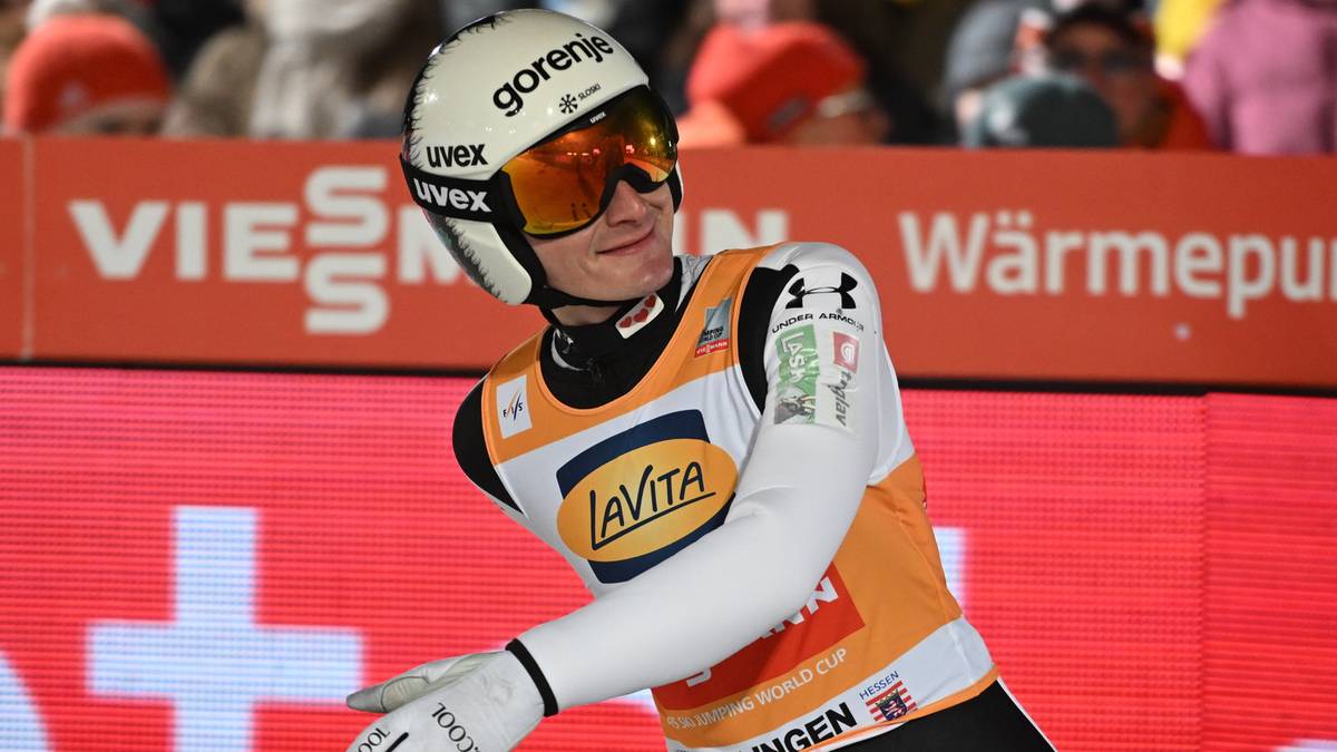 Skoczek narciarski w białym kasku z goglami na głowie i w białych rękawiczkach, z uśmiechem na twarzy, stoi na tle czerwonej tablicy z napisem "Willingen".