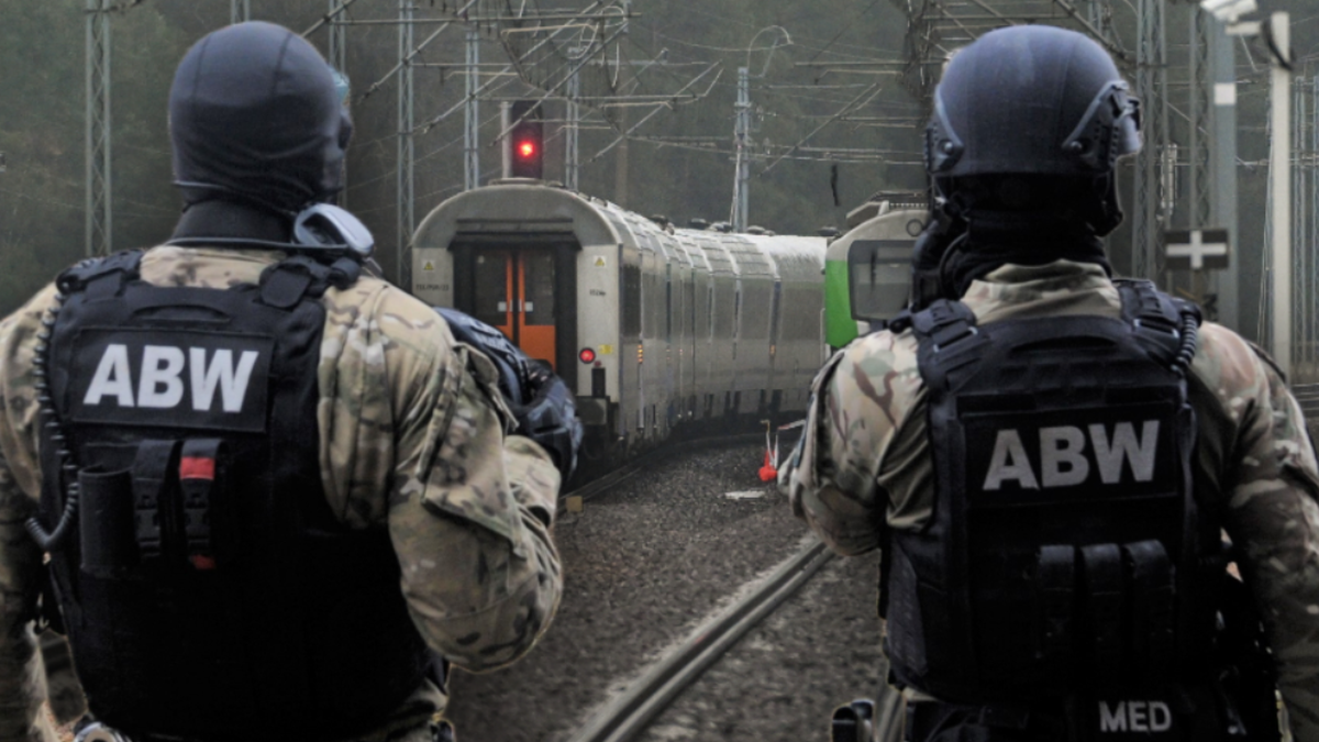 Dwóch funkcjonariuszy ABW w kamizelkach taktycznych z napisem ABW stoi tyłem do widza i obserwuje tory kolejowe, na których stoi pociąg.