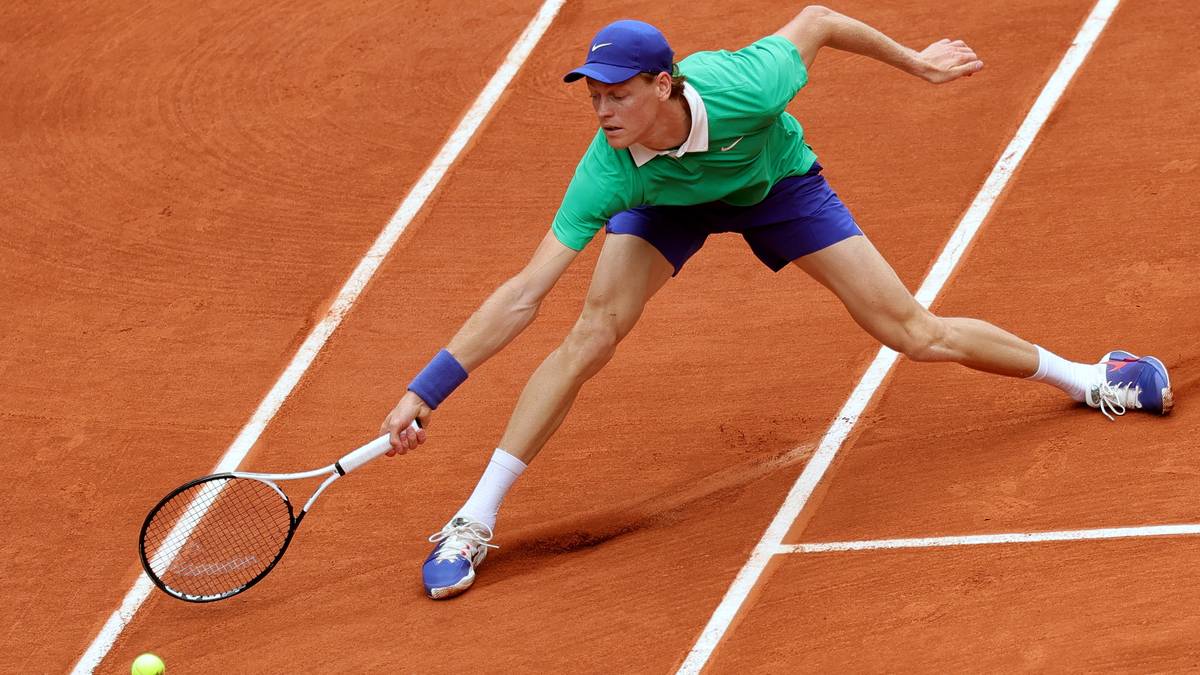 Janik Sinner w akcji na korcie ziemnym, w zielonej koszulce i granatowych spodenkach, wykonuje zamaszysty forhend z lewej strony.
