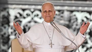 Papież Leon XIV w białych szatach przemawia do mikrofonu z rozłożonymi rękami.