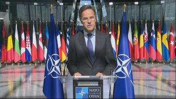 Mężczyzna w garniturze i krawacie stoi za m&oacute;wnicą z logo NATO, na tle flag państwowych.