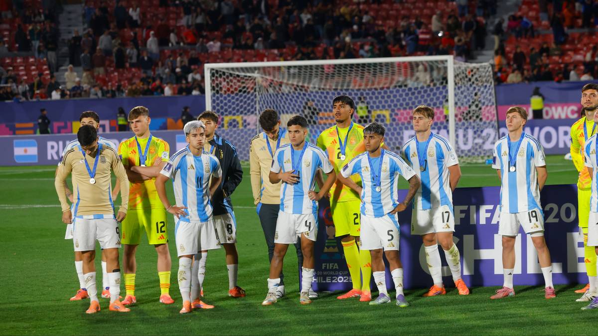 Grupa piłkarzy w niebiesko-białych strojach stoi na boisku, otrzymując medale po meczu. Na drugim planie widać trybuny wypełnione widzami oraz bramkę.