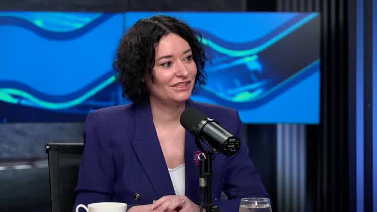 Kobieta z kr&oacute;tkimi włosami w granatowej marynarce m&oacute;wi do mikrofonu. Na stole stoi filiżanka i szklanka. W tle ekran z niebieskim wzorem.