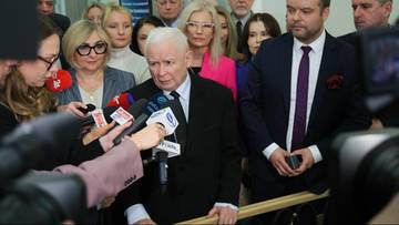 Jarosław Kaczyński przemawia do dziennikarzy otoczony przez grupę os&oacute;b.
