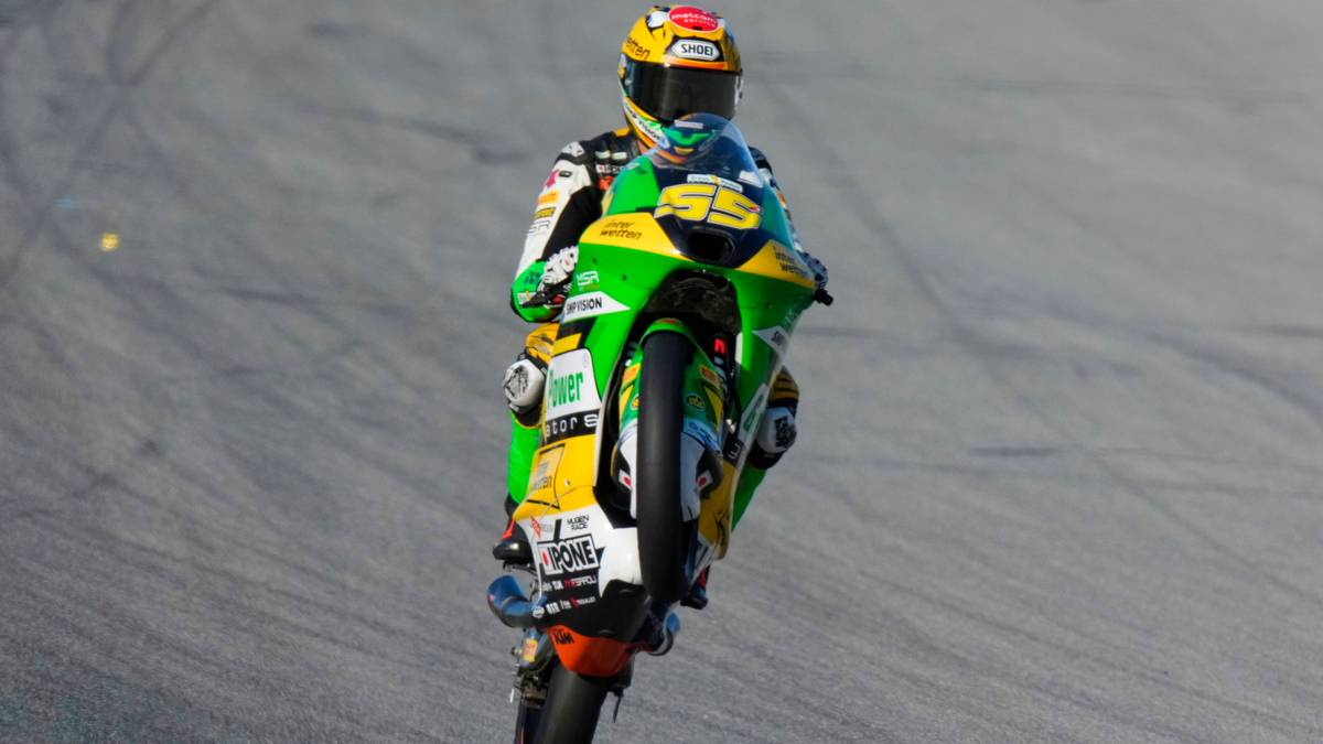 Motocyklista w zielono-żółtym stroju wyścigowym wykonuje jazdę na jednym kole na torze wyścigowym.