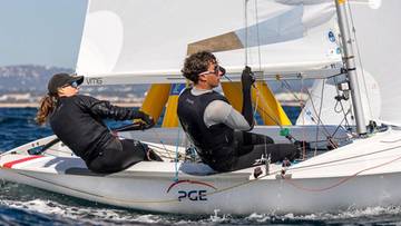 Dwoje ludzi na żagl&oacute;wce, jeden steruje, drugi obsługuje żagiel. Osoby ubrane w stroje sportowe, na wodzie.