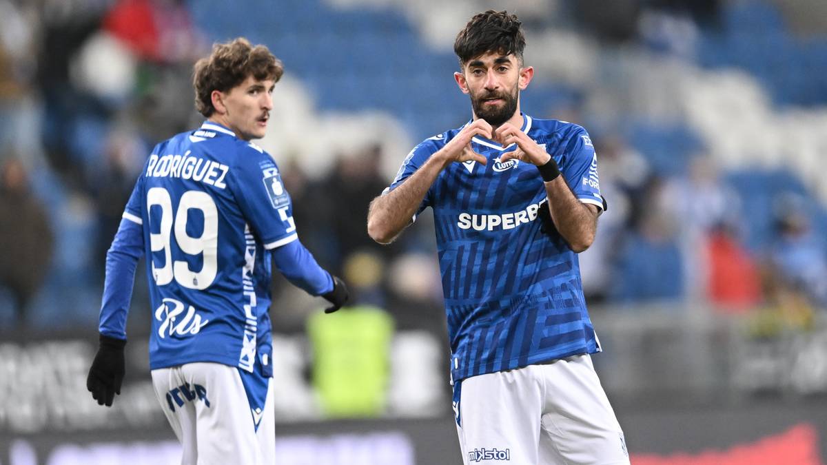 Dwóch piłkarzy w niebieskich koszulkach na boisku piłkarskim, jeden z nich pokazuje gest serca dłońmi.