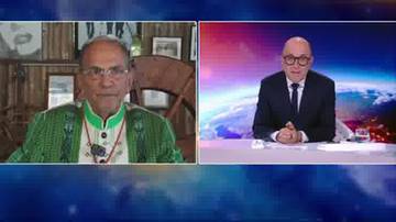 Dwie połowy obrazu. Na lewej, mężczyzna w okularach i zielonej, wzorzystej kurtce siedzi przed drewnianą ścianą. Na prawej, mężczyzna w okularach, garniturze i krawacie siedzi przy stole w studio telewizyjnym z widokiem na Ziemię.