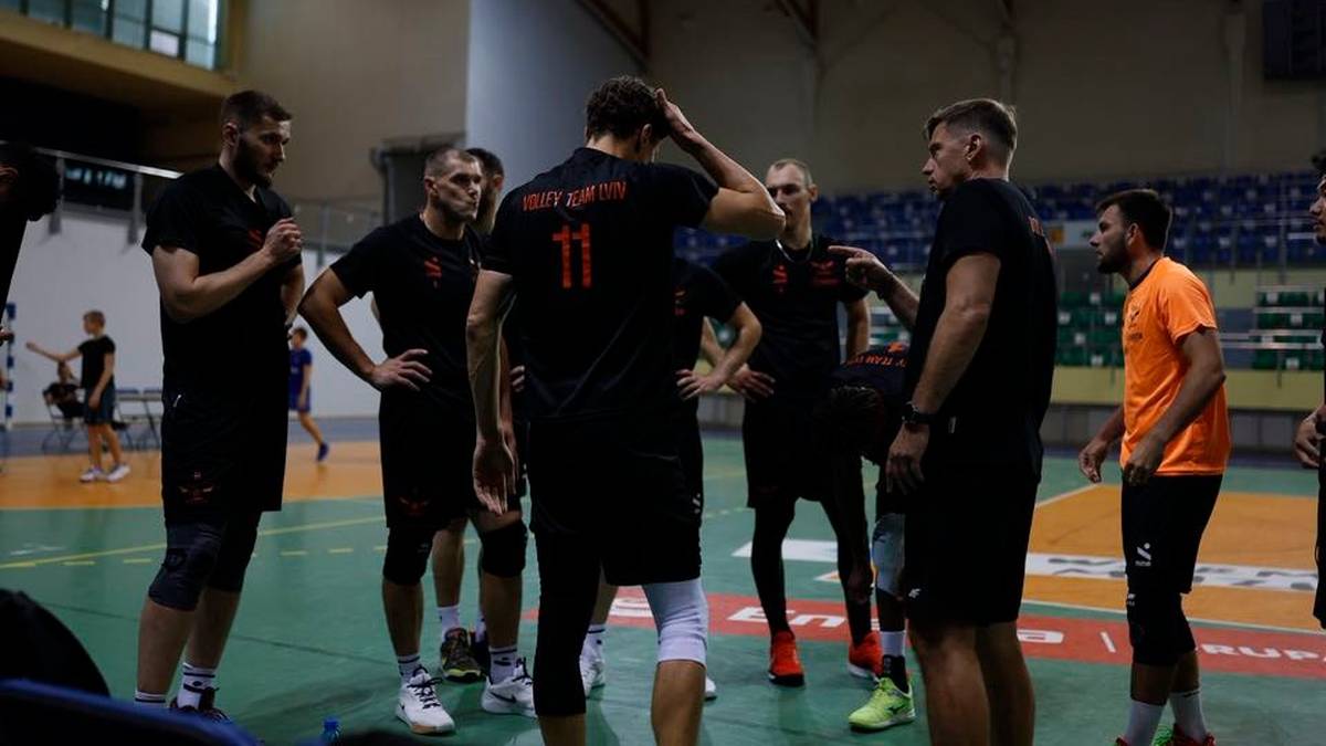 Grupa siatkarzy w czarnych strojach stoi w kręgu na boisku, rozmawiając. Jeden z nich trzyma rękę na głowie.