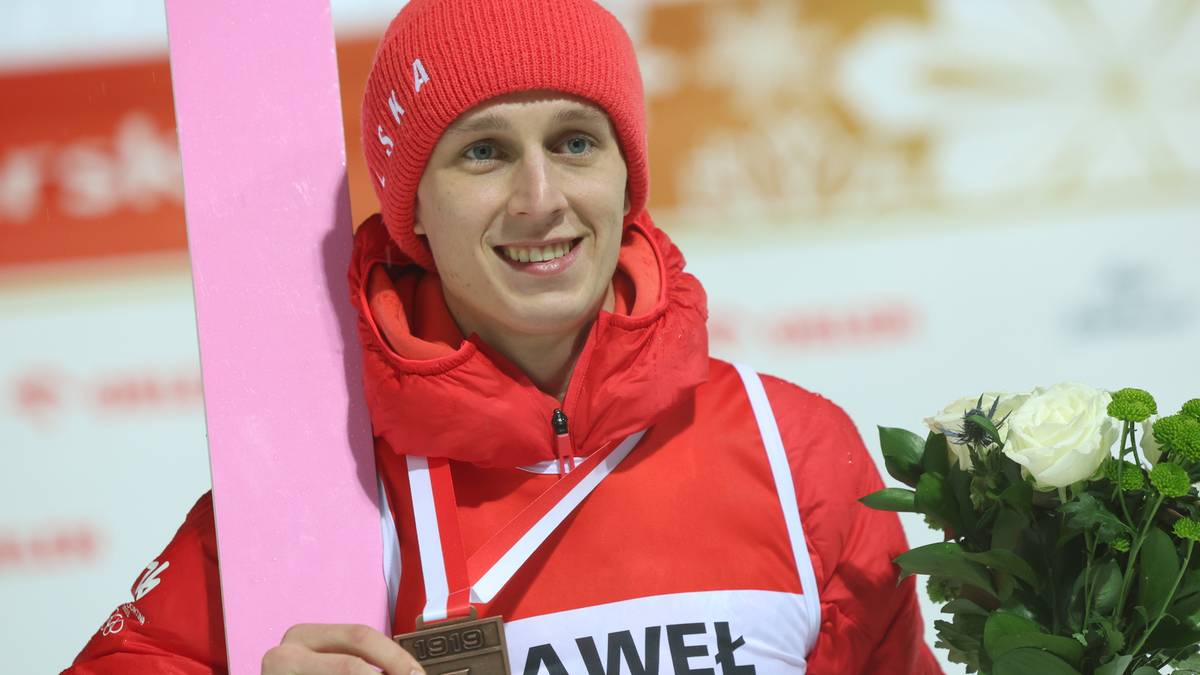 Mężczyzna w czerwonej czapce i kurtce z białymi elementami, trzymający medal i bukiet białych r&oacute;ż.