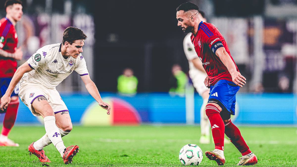 Dw&oacute;ch piłkarzy walczy o piłkę na zielonym boisku podczas meczu piłki nożnej, jeden w białej koszulce, drugi w czerwonej z niebieskimi elementami.