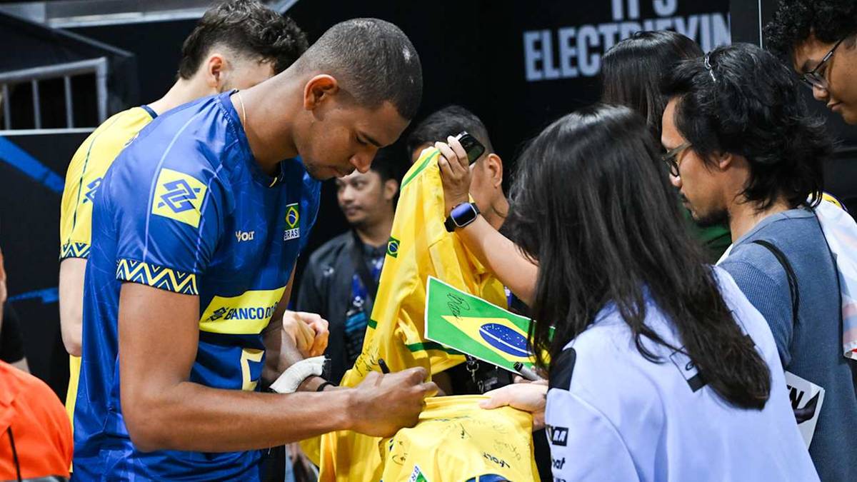 Siatkarz w niebieskiej koszulce reprezentacji Brazylii bierze do ręki flagę Brazylii od kibiców.