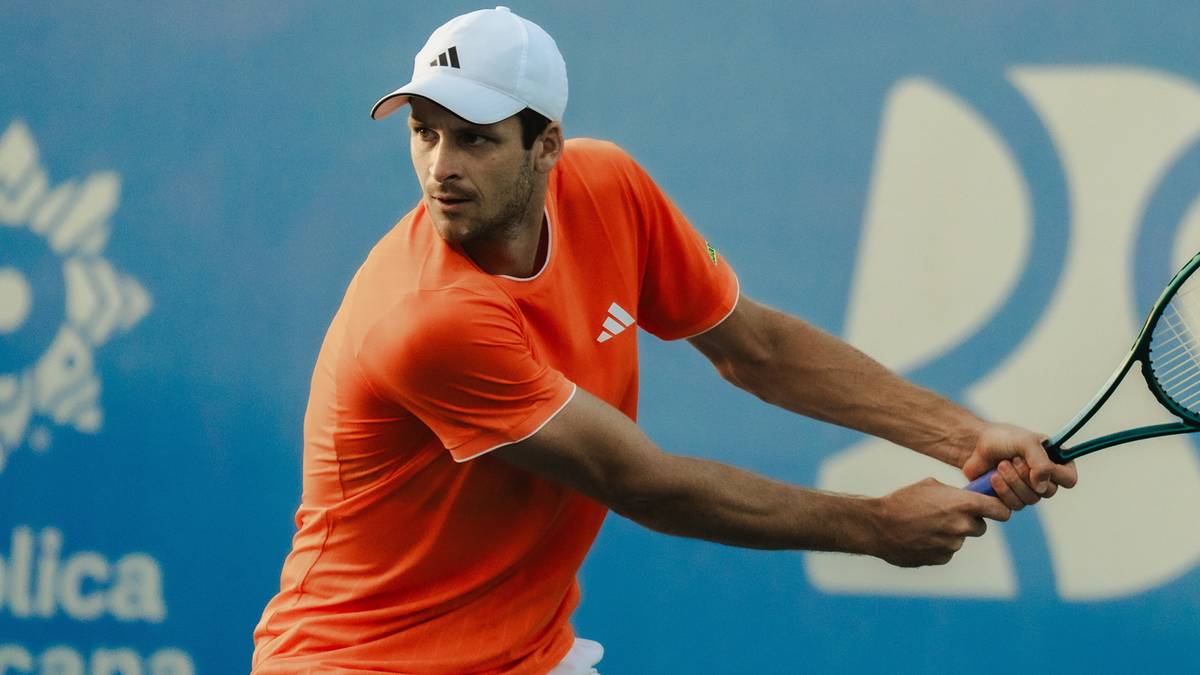 Tenisista w pomarańczowej koszulce i białej czapce uderza piłkę rakietą podczas meczu na korcie tenisowym.