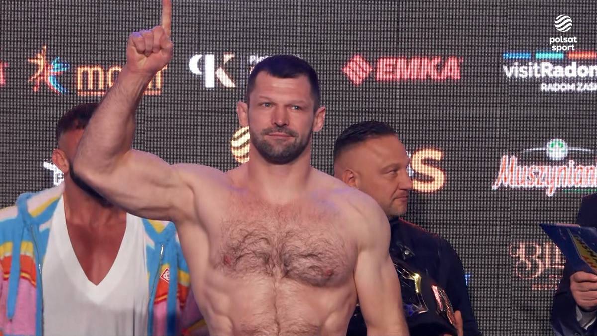 Mężczyzna z uniesionym palcem wskazującym, prezentujący muskulaturę na tle banerów.