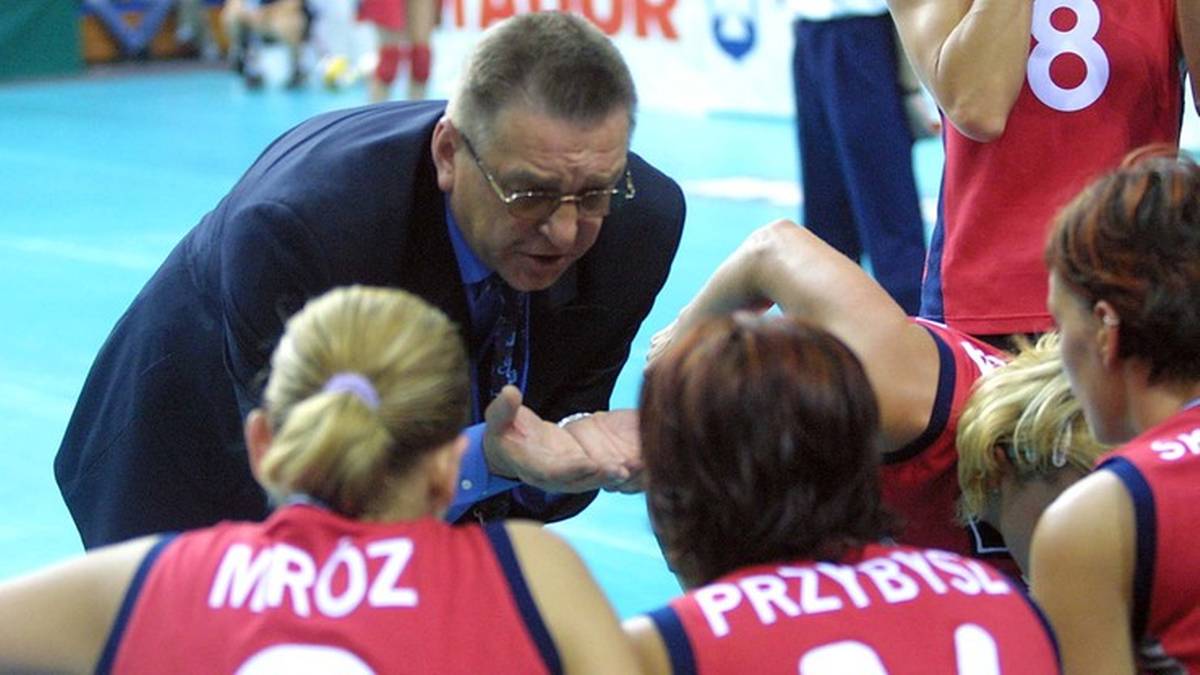 Trener w okularach i ciemnym garniturze pochyla się nad kilkoma zawodniczkami siatk&oacute;wki w czerwonych strojach, udzielając im wskaz&oacute;wek.