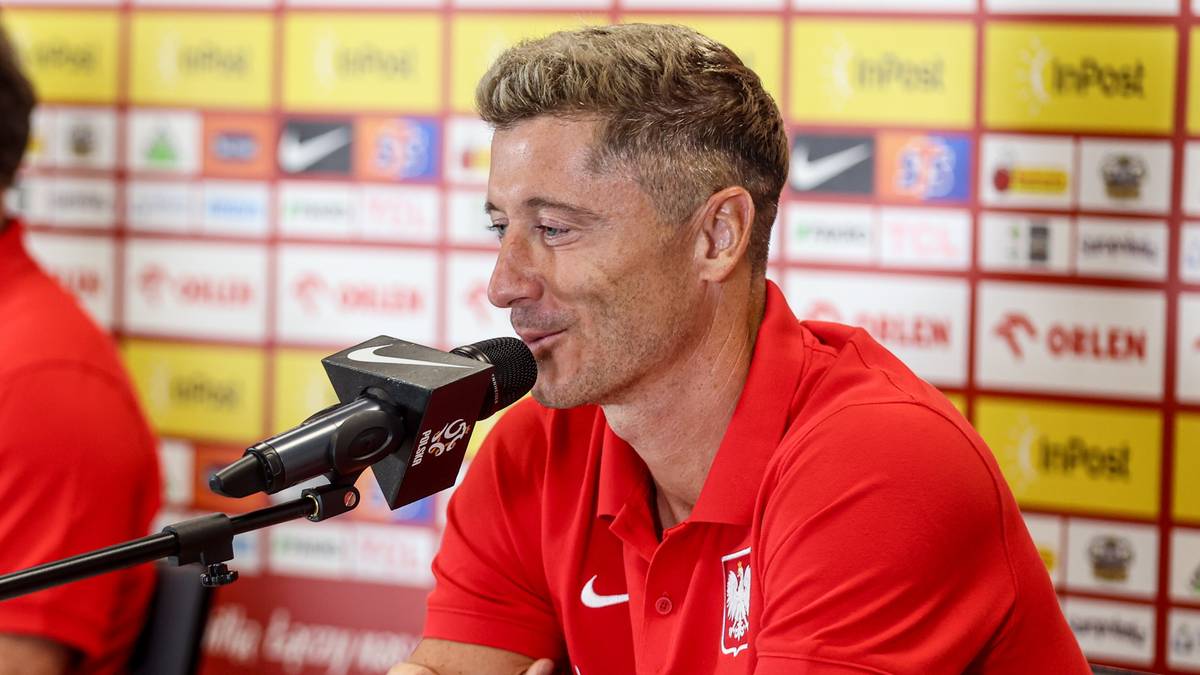 Robert Lewandowski w czerwonej koszulce z polskim godłem, przemawiający do mikrofonu.