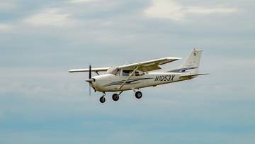 Biały samolot Cessna leci na tle niebieskiego, zachmurzonego nieba.