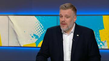 Mężczyzna w średnim wieku z brodą i kr&oacute;tko przystrzyżonymi włosami, ubrany w ciemny garnitur i białą koszulę, m&oacute;wi do kamery. W tle znajduje się nowoczesna scenografia telewizyjna z kolorowymi, geometrycznymi elementami.